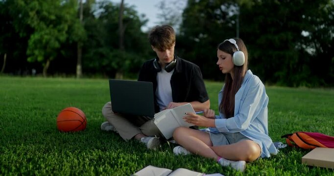Focused teenage girl asks for help from her boyfriend classmate while self-development and doing homework on the green lawn in the park