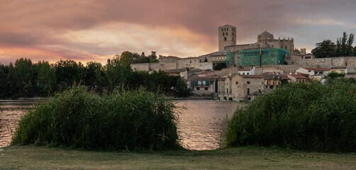 Photograph of Zamora, highlighting the Cathedral, seen from the other side of the Duero River at dusk, with the setting sun bathing the scene in golden and orange hues. Zamora, Spain