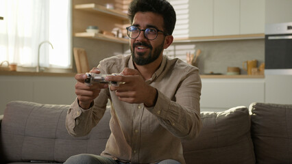 Indian Arabian man ethnic guy have fun on sofa couch play online video game smart TV console virtual driving car use wireless controller entertainment in home kitchen male gamer playing with joystick