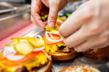 Close-up of hands placing pickles on a freshly made burger with lettuce, tomato, and cheese. Perfect for food preparation, culinary arts, and fast food themes. High-quality, appetizing, and vibrant.