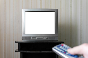 Old television with blank white screen and hand holding remote control in foreground. TV is placed...