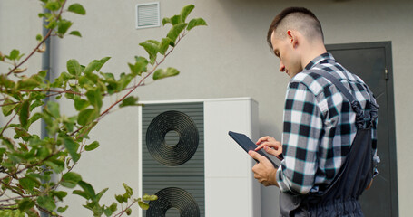 A technician uses a tablet to adjust settings on an external heat pump unit outside a home.