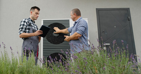 A technician consults with a homeowner at an external heat pump unit, discussing maintenance...