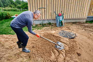 Builder compacts sand near septic tank manhole cover, patting its surface with shovel.