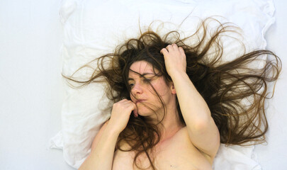 sexy nude woman with disheveled hair lies on a white pillow, her expression distant and reflective....