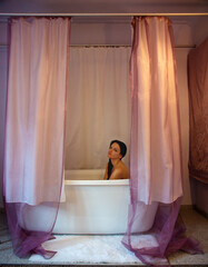 Sexy Young woman sits in white hotel bathtub surrounded by soft pink and purple curtains, creating an intimate, serene atmosphere, thoughtful expression, contemplative mood