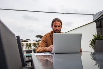 young athletic guy, in smart working mode, is busy with his laptop with focus and dedication, while sitting on the table in his home garden with swimming pool