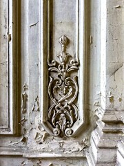 A close-up of an intricate decorative carving on the peeling wall of a decaying abandoned palace. The elaborate design features delicate scrollwork and floral patterns, hinting at the former grandeur