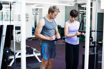 Coach, woman and clipboard with weightlifting machine, assessment and progress at gym. People, man and personal trainer with checklist with workout, fitness and exercise with notes at wellness club