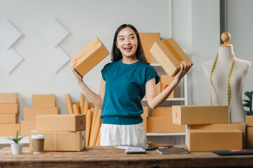 Happy Small Business Owner: A young Asian woman with a bright smile holds up two cardboard boxes in...