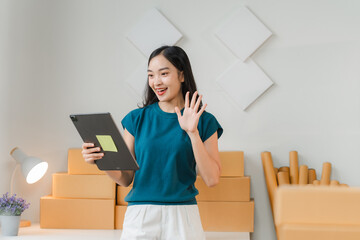 Smiling Businesswoman on Video Call: A young Asian businesswoman with a warm smile waves hello during a video call, surrounded by stacked cardboard boxes, showcasing the busy and dynamic world of entr