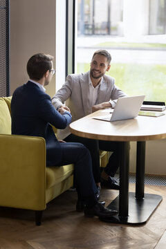 Happy business partners men giving handshakes with respect, recognition, thanking for good job, meeting in office cafe, sitting at laptop. Businessman hiring candidate after successful interview