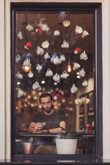 Man shop location- drinking coffee- looking out of window. Through window view of positive thoughtful young male enjoying cup of hot coffee while resting in cozy cafe