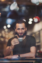Man shop location- drinking coffee- looking out of window. Through window view of positive thoughtful young male enjoying cup of hot coffee while resting in cozy cafe