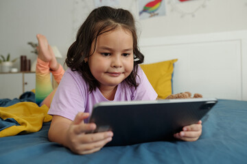 Little girl lying in her bed and watching cartoons on digital tablet