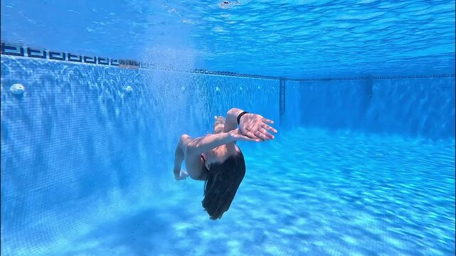 A girl jumps headfirst into the pool and swims mermaid style under the water with a bright, clear blue background, approaches and smiles
