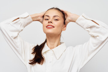 Naklejka premium Portrait of a confident young woman with a charming smile, wearing a stylish white shirt, hands raised behind her head, set against a light grey background