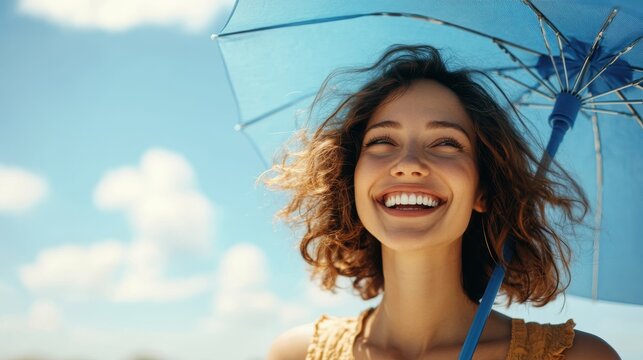 A young woman laughs joyously while using a blue umbrella on a sunny day, embodying positivity and carefree spirit in an outdoor setting.