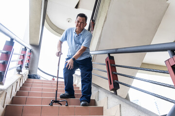 Elderly Asian man with extreme knee pain holding walking stick as support to go down staircase