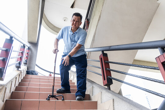 Elderly Asian man with extreme knee pain holding walking stick as support to go down staircase - Powered by Adobe