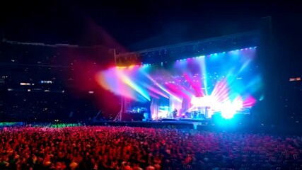 A packed concert arena filled with an energetic audience, with bright lighting effects illuminating the stage in a vibrant display of music and motion