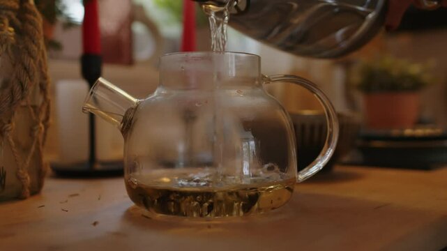 Closeup of anonymous person pouring hot water from kettle into glass teapot with tea leaves to brew up black tea at domestic kitchen