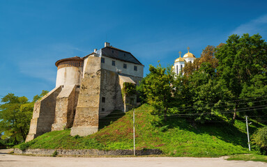 Fototapeta premium Ancient Ostroh Castle with its round tower stands on a green hill, alongside golden-domed Orthodox church. The historic fortress in Rivne region, Ukraine, basks in bright sunlight.