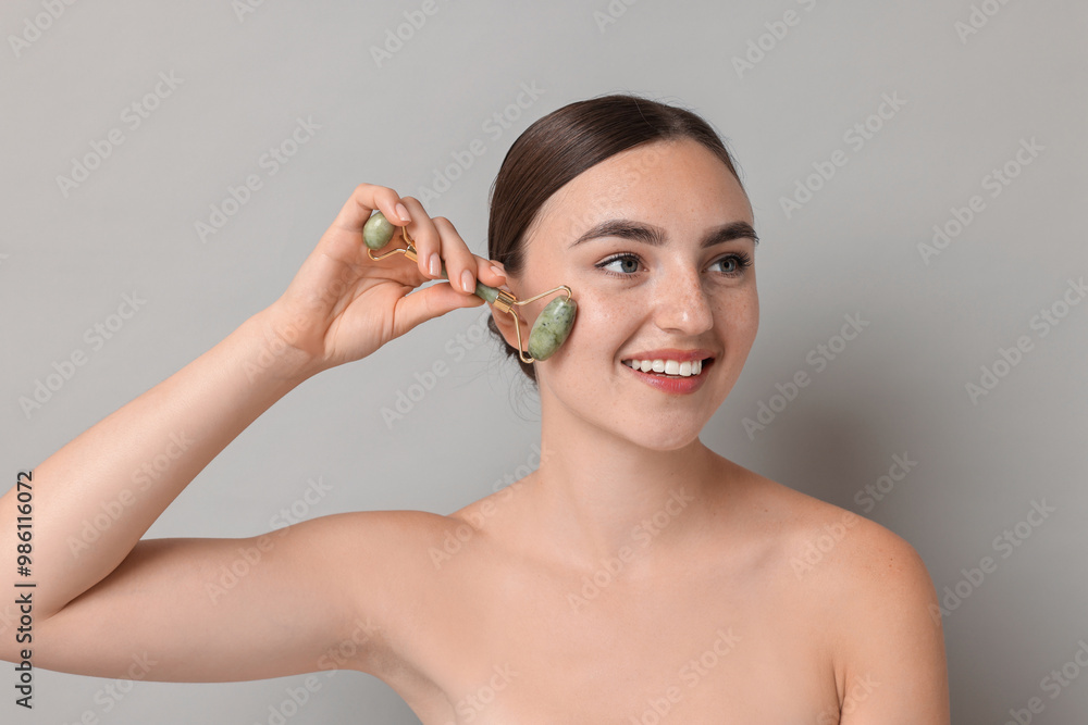 Wall mural Beautiful young woman doing facial massage with roller on grey background