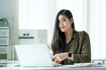 Professional young woman with long dark hair is sitting at a bright, modern workspace, holding a smartphone while looking confidently at the camera.