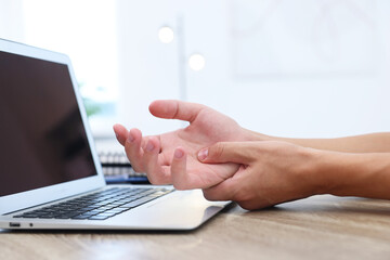Man suffering from pain in wrist while working on laptop at table indoors, closeup. Carpal tunnel...