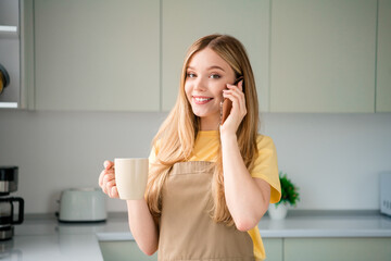 Fototapeta premium Photo of attractive teen woman drink coffee talk phone dressed apron enjoy preparing delicious food kitchen indoors
