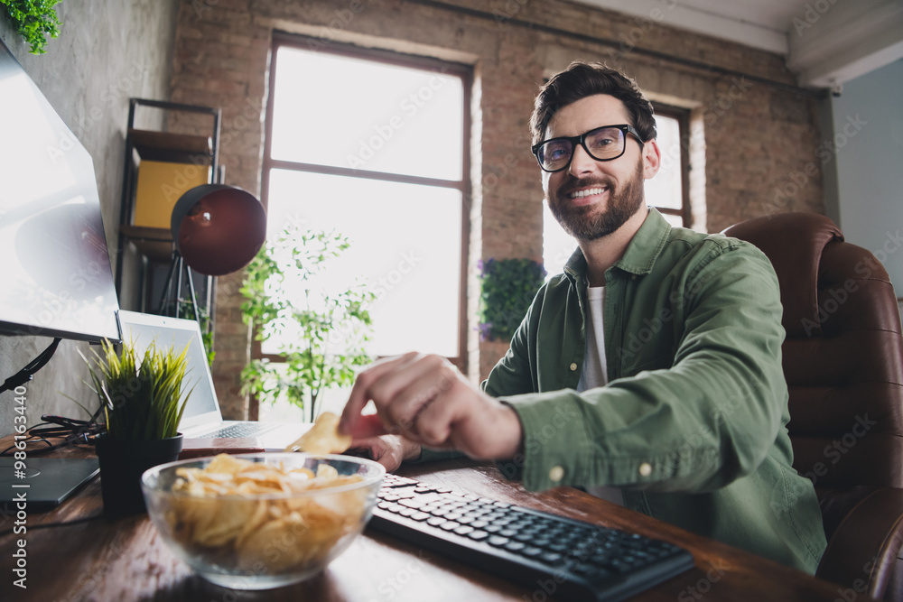 Poster Portrait of professional hacker young man eat chips computer desk loft interior office indoors