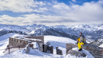 Men in snowboard gear sitting on a stone wall and looking in to distance, snowy mountains in background, winter sport, Montgenevre, France, Alps