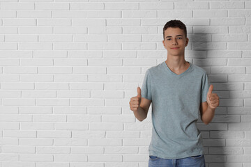 Teenage boy wearing grey t-shirt and showing thumbs up near white brick wall, space for text