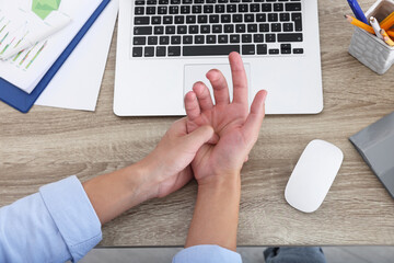 Man suffering from pain in wrist while working on laptop at wooden table, closeup, top view. Carpal...