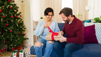 Latin woman is opening a present given by her boyfriend while sitting on the sofa in a cozy decorated living room