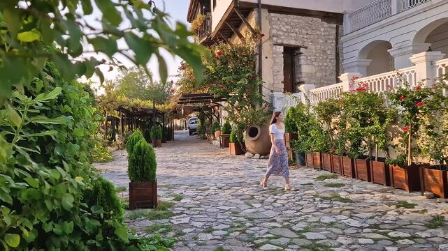 Beautiful young woman walking on the cobblestone narrow streets of Old Nessebar town admiring the coastline traditional buildings surrounded by greenery. Summer vacation travel in Bulgaria