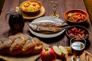 Challah, honey, apple and pomegranate and wine and chelnt and fish on the table with menorah and burning candles in honor of Rosh Hashanah