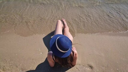 Rear view young woman wearing a blue sunhat sitting on the beach with legs stretched towards the sea. The clear water is gently lapping her feet. Serene and relaxing summer holiday scene - Powered by Adobe