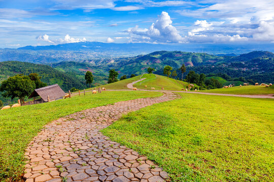 A scenic view of a stone pathway leading through green pastures, rolling hills, and a traditional farmhouse. The beautiful mountainous landscape and distant clouds create a peaceful, idyllic scene.

