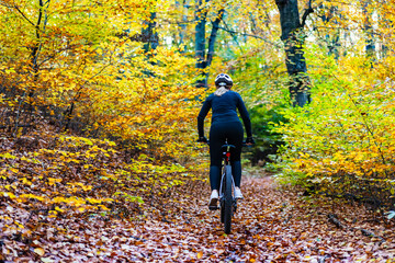 Fototapeta premium Woman wearing black sporty jacket, black cycling pants and white bike helmet riding bicycle in forest in autumn scenery. Back view.