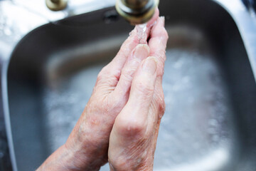 Close-up of elderly lady washing her hands under a running tap against a silver shade sink 