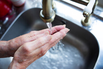 Close-up of elderly lady washing her hands under a running tap against a silver shade sink 