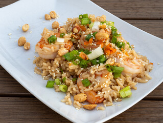 Top view of a shrimp fried rice dish on white plate, topped with peanuts, scallions, and sesame seeds on wooden table.