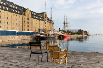 A tranquil waterside scene is shown, with a wooden dock and two chairs facing the water. There are two old boats docked and a large building in the background.