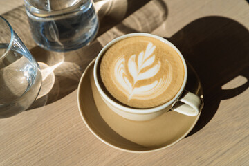 Close-up of cappuccino in beige ceramic cup, a jar and a glass of water on a wooden table in the sunlight