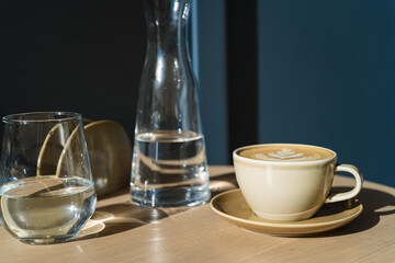 Cappuccino in beige ceramic cup, a jar and a glass of water on a wooden table against dark blue wall in the sunlight