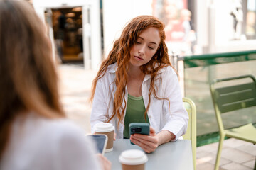Young Woman Relaxes Outdoors, Absorbed in Her Smartphone While Sipping Coffee on Sunny Day