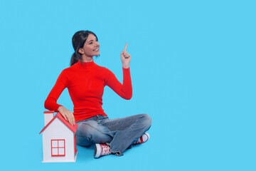Confident Woman Sharing Bright Idea While Sitting Next to  Model House Against a Blue Backdrop