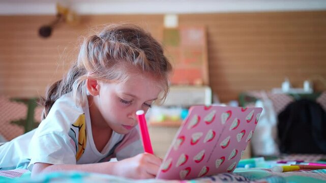 Small child lies on bed in room drawing with a pink marker in a pink notebook 